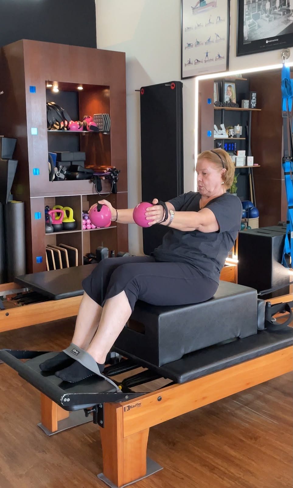 Alumna adulta mayor sentada sobre la Sitting Box del Reformer con una pelota rosada en las manos